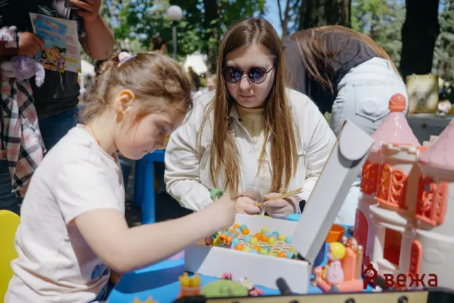 Child wearing sunglasses concentrates on threading colorful beads at an outdoor bead craft table; a second child watches nearby.