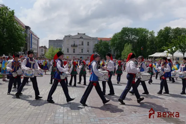 A street parade with people in colorful traditional costumes marching and playing drums in a sunny city square.