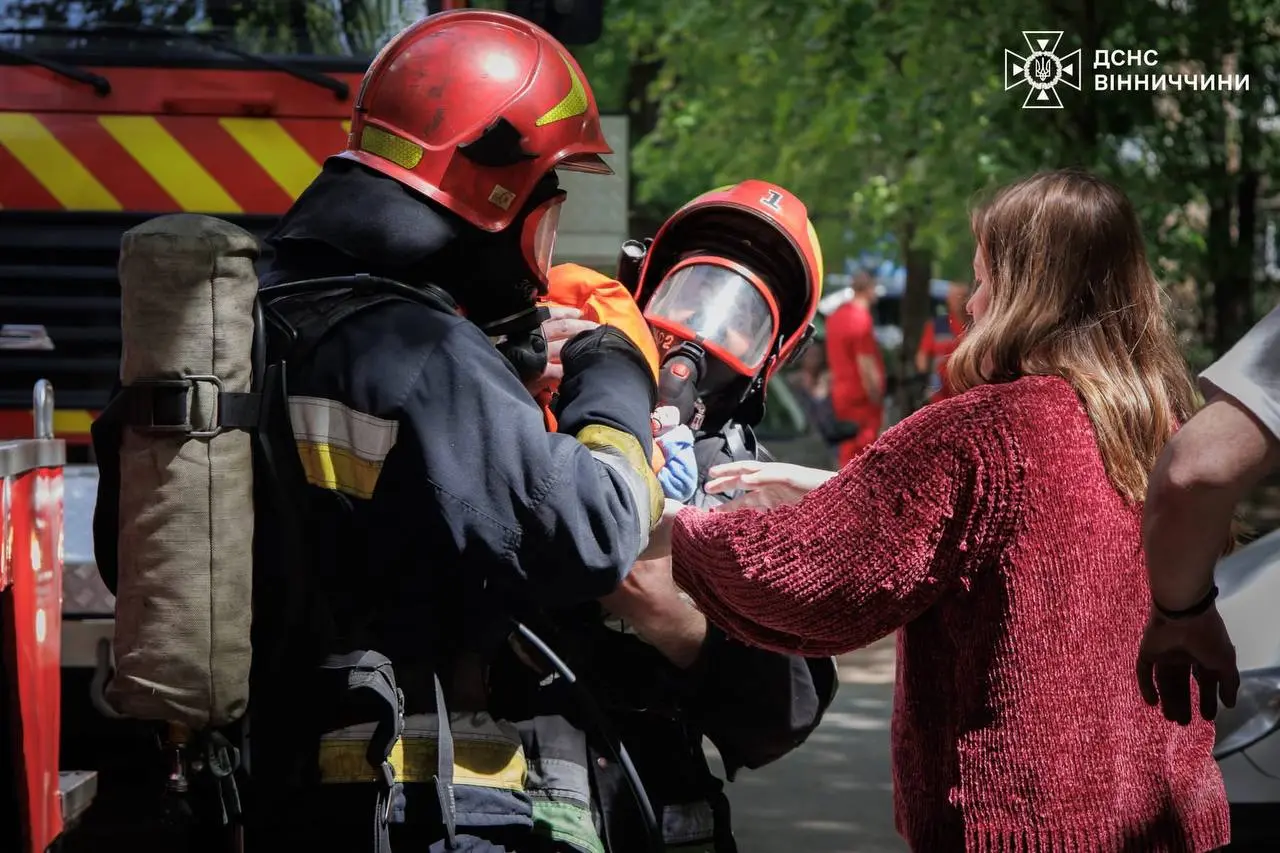 Two firefighters in red helmets help a woman with a safety gesture during a public safety demonstration beside a fire truck, Vinnytsia region logo visible on the top right.