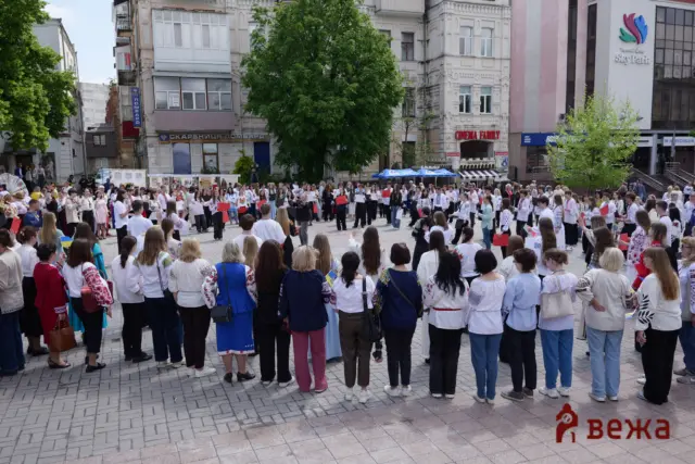 Large crowd of people wearing embroidered traditional Ukrainian clothing gathers in a city plaza, forming a circle around the center while modern buildings and a big tree loom in the background, with some holding red and white signs.
