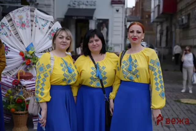 Three women in matching yellow embroidered blouses and blue skirts pose together at a street festival.