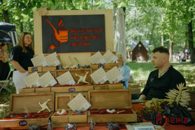 Park market stall selling handmade wooden figurines; display boxes with price tags and a large black sign with red Ukrainian text, trees in background.