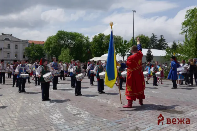 Group of Ukrainian folk drummers in traditional outfits marching, with a blue–yellow flag in a town square outdoor parade.