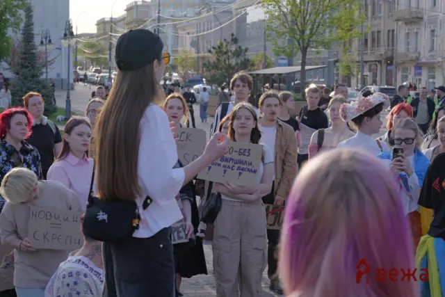 Crowd of protesters on a city street listening to a speaker, holding cardboard signs with Cyrillic text in daylight.