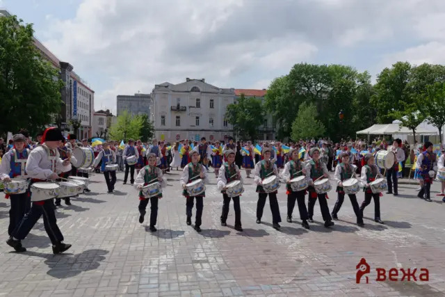 Drummers in colorful Ukrainian folk costumes marching in a city square, waving small blue-yellow flags amid a crowd and trees.
