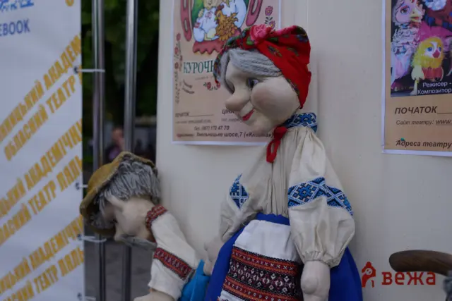 Handmade fabric puppet wearing a red floral headscarf and embroidered dress stands beside posters on an outdoor wall.