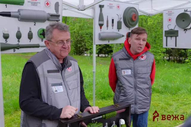 Two men in gray vests stand beside an outdoor display of emergency tools and medical gear mounted on white panels, with greenery in the background.