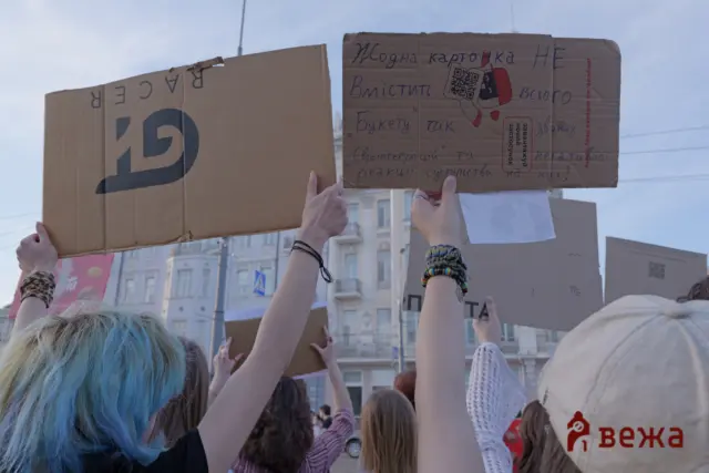 Crowd of protesters raising handmade cardboard signs above a city street; several raised fists visible.