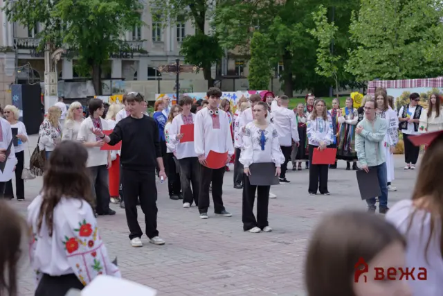 Crowd of people in traditional Ukrainian clothes holding red folders at an outdoor festival in a town square with green trees behind them.