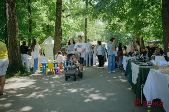 Shoppers stroll through an outdoor market in a leafy park, with tents, vendors, and a baby stroller in the foreground.
