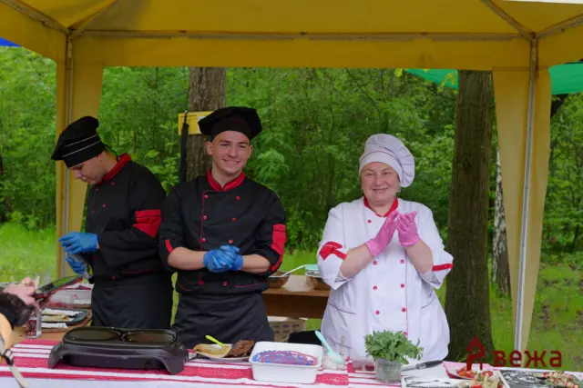 Three chefs in a yellow outdoor tent preparing food at a table with cutting boards and herbs, smiling at a cooking event.