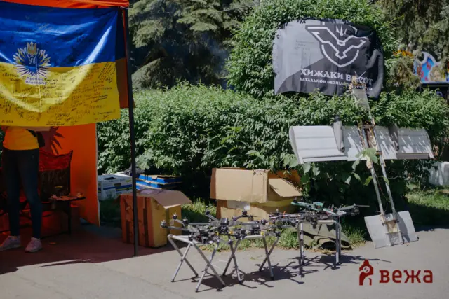 Outdoor booth under an orange canopy with a signed blue-and-yellow Ukrainian flag, cardboard boxes, and metal stands near greenery.