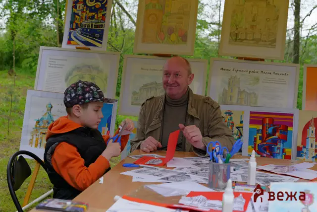 Man and child crafting red paper shapes at an outdoor art booth, with framed colorful illustrations and drawings in the background.