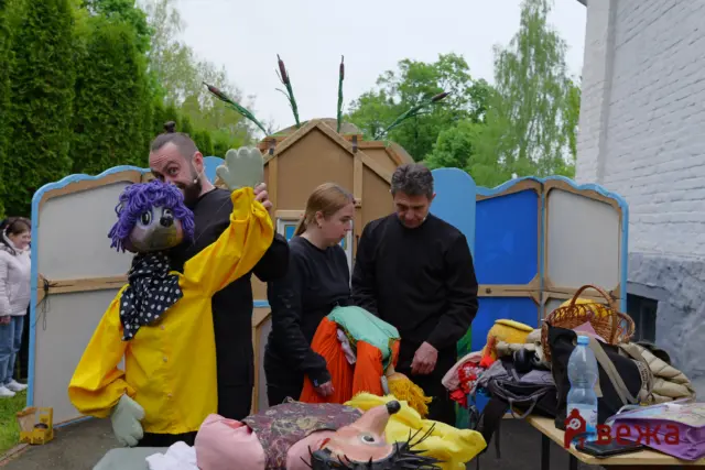 People in black costumes arranging colorful puppets and props at an outdoor puppet show setup, with a bright yellow puppet suit and a large purple puppet head in the foreground.