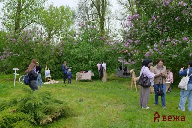 People at an outdoor garden market among blooming lilac bushes and green trees.