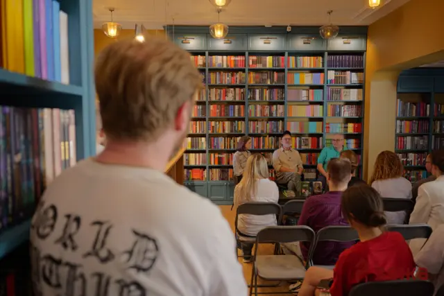 Panel discussion in a bookstore: speakers seated at the front with an audience, bookshelves filled with colorful titles behind them.