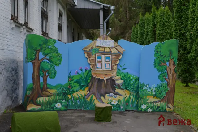Colorful painted garden scene on a blue panel featuring a small wooden cabin, trees, and flowers beside a white brick building.