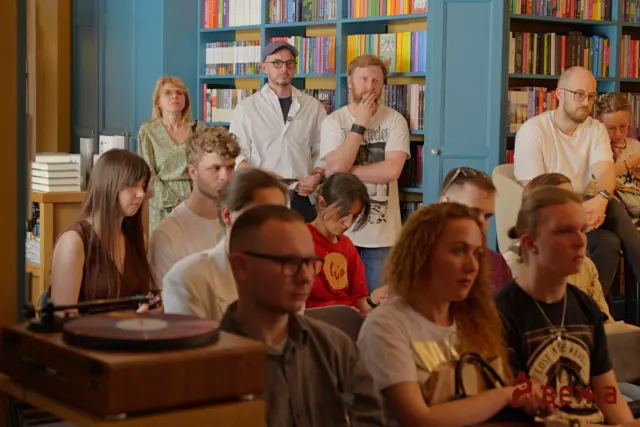 Group of people at a bookshop event, standing and seated, with blue bookshelves behind them.