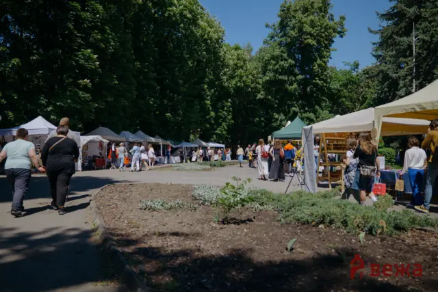 Outdoor market scene with white tents along a tree-lined path and people browsing stalls on a sunny day.