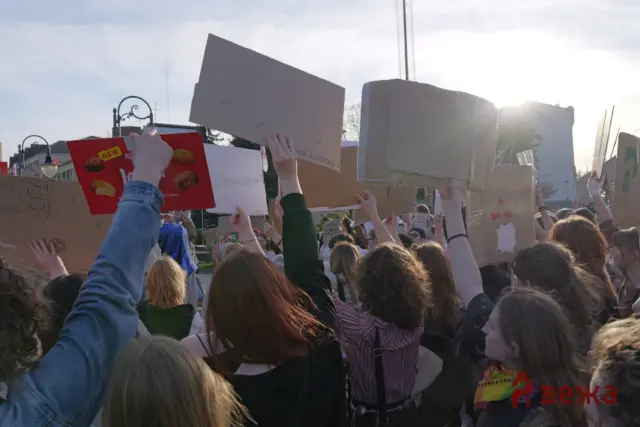 Crowd of protesters holding cardboard signs during a demonstration, with sun glare in the background.