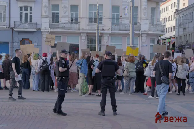 Crowd of protesters holding cardboard signs in a city square with ornate white buildings behind them.