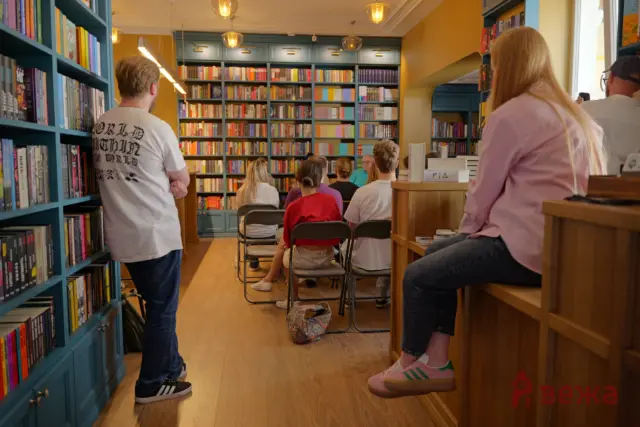 Audience seated in a bookstore reading area, facing a wall of colorful books on blue shelves with warm lighting above.