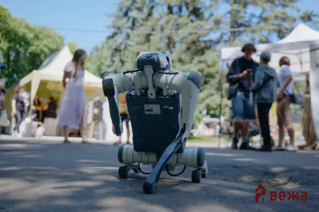 Humanoid robot on wheels at an outdoor event with people and tents in the blurred background.