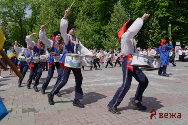 A group of drummers in colorful folk costumes marching in a park-style parade, raising drumsticks mid‑beat.