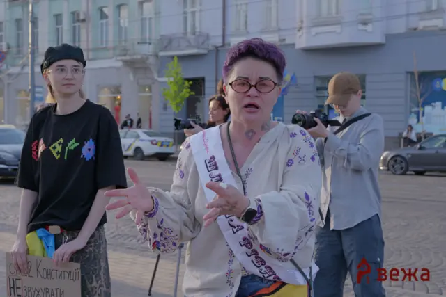 Group of protesters in a city square, woman with purple hair and sash speaking, others hold signs in Ukrainian behind her