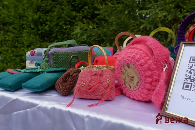 Colorful handmade crafts on a table: pink fuzzy slippers, woven baskets, and other textiles outdoors with a QR code frame nearby.
