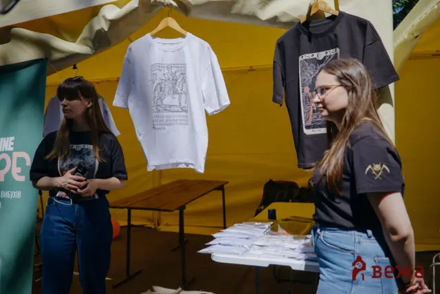 Two women stand at a booth displaying graphic T‑shirts on hangers, with a yellow tent backdrop and a table of papers and merchandise.