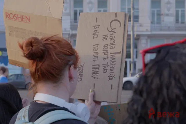 Person with red hair seen from behind holds a large cardboard sign with handwritten text at a street protest.