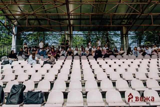 Group of students seated on light pink stadium seats under a rusted metal roof in an outdoor arena, with trees in the background.