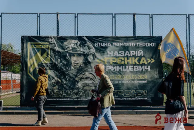 Outdoor event with a large military banner mounted on a chain-link fence and people walking by.