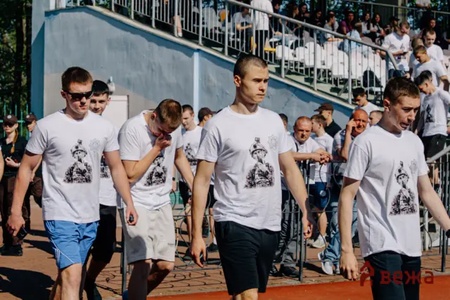 Group of men in white t-shirts with graphics walking along a fenced outdoor area, with spectators on bleachers behind them.