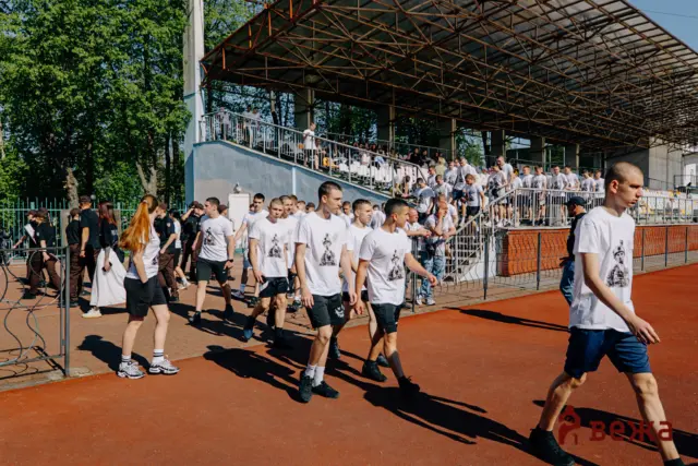 Large group of people in white T-shirts walk along a red track outside a stadium with spectators in the stands.