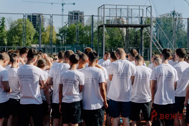 Group of people in white T-shirts standing with their backs to the camera on a sports field, fence and buildings behind them.