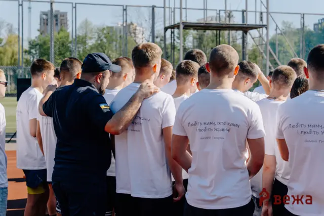 Men in white T-shirts huddle on an outdoor sports field, a coach with his arm around players.