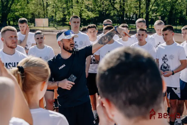 Instructor with a tattooed arm points and speaks to a group of young men in white shirts outdoors during training in a sunny park.