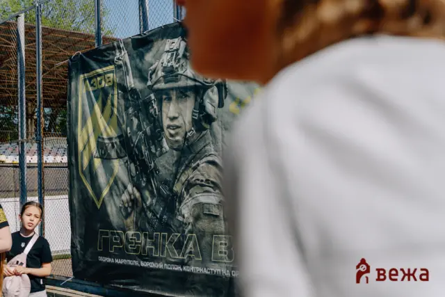 Outdoor scene with a large banner showing a soldier image behind a chain-link fence, two children standing to the left, blurred foreground on the right.