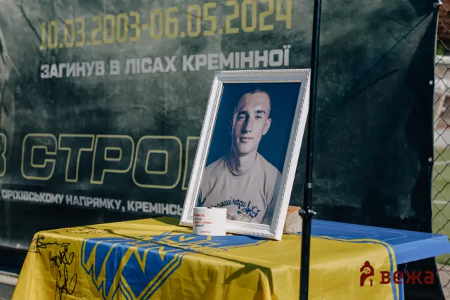 Framed portrait of a man on a table covered with blue-yellow cloth, placed at a memorial display with a large Cyrillic banner behind.
