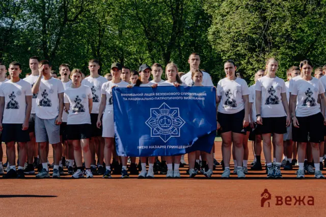 Group of young people wearing white T-shirts holding a blue banner outdoors on a running track, with green trees in the background.