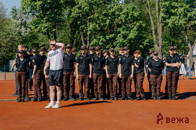 Group of uniformed cadets standing in formation on an outdoor track, with a man in a white shirt in front shading his eyes from the sun, trees in the background.