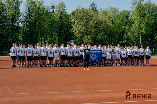 Large group of people in white shirts standing on a red running track outdoors, front row holding a blue banner with trees behind them.