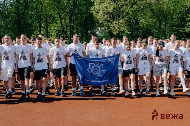 Group of young men in white shirts marching on a running track, holding a blue banner with a crest, trees in the background.