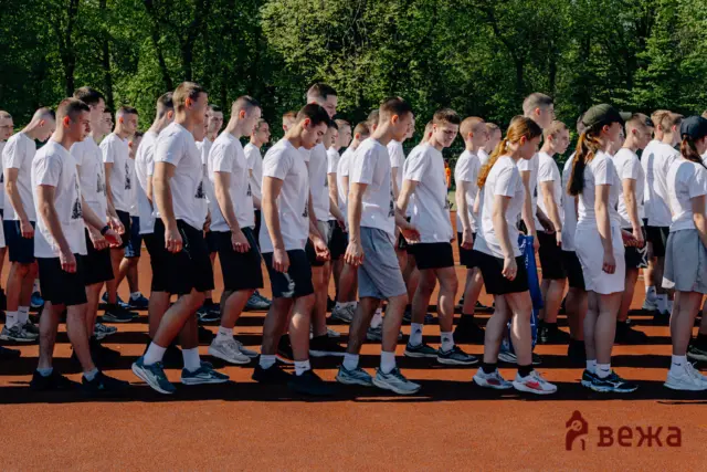 Line of young people in white shirts walking on a track outdoors, trees in the background.