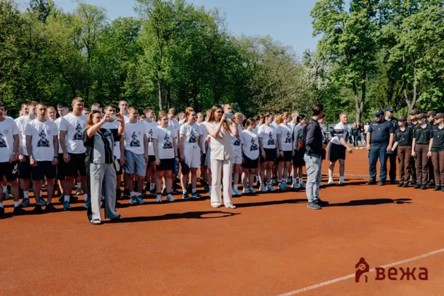 Group of people in white shirts standing on a red outdoor track, some taking photos, with trees in the background at a sunny event.