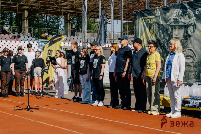 Group of people standing on an outdoor stage with microphones, banners, and spectators in stadium seating behind them.