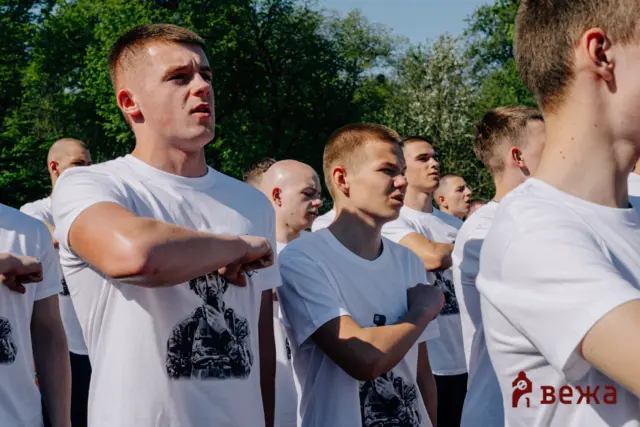 Group of young men in white T-shirts standing with arms crossed outdoors, in a formation among trees.