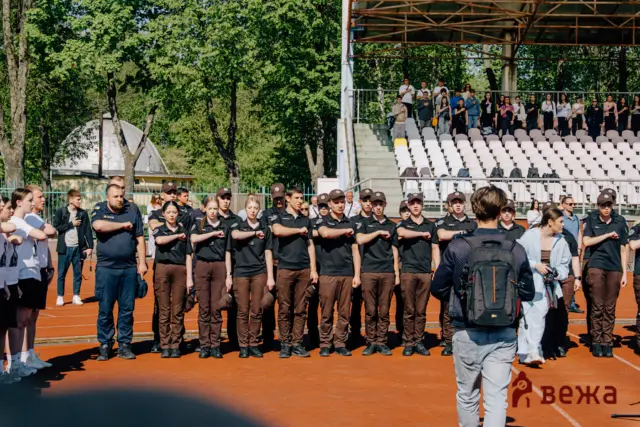 Group of uniformed individuals standing in formation on a running track with a bleacher audience behind them on a sunny day.